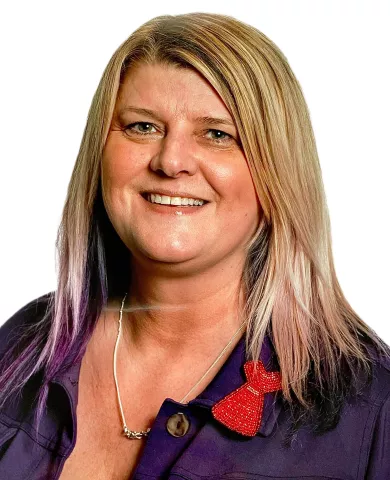 A smiling blond woman with purple tips on her hair and a red dress pin on her lapel.