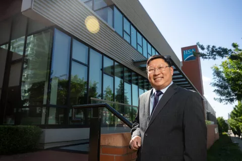 A smiling Kane Tse wearing a charcoal suit in front of the HSA office.