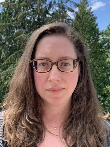 A cropped image of white woman with brown glasses and long brown hair, standing in front of trees and sky.