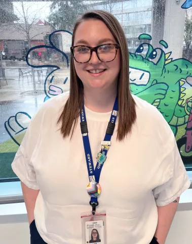 A smiling woman in a white top, wearing large glasses and an HSA lanyard, standing in front of a painted window.