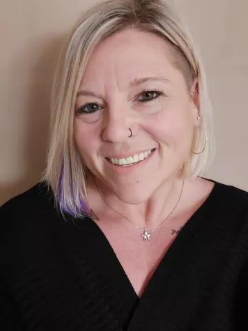 A smiling worman with very blonde hair with purple tips, wearing a black top and smiling at the camera,