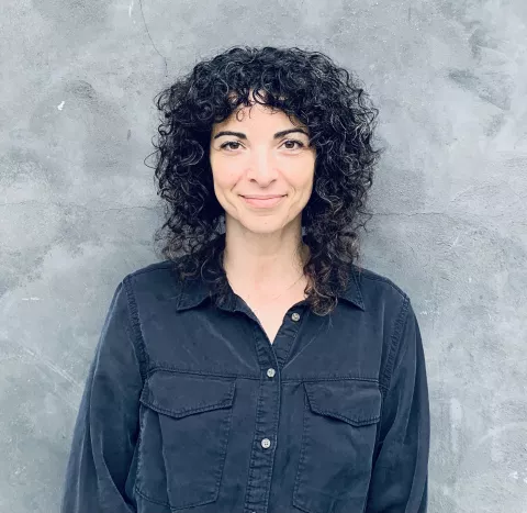 A woman with dark, curly, shoulder-length hair, wearing a dark blue button down and standing in front of a neutral backdrop.