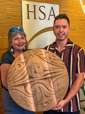 Two people, standing in front of HSA's lobby, holding a beautiful round cedar wood carving.