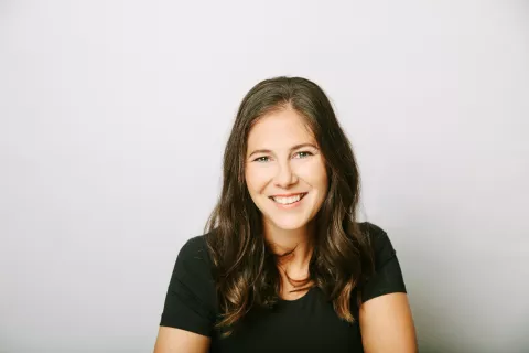 A smiling woman with long brown hair wearing a black t-shirt; only her head and shoulders are visible in front of a plain gray background.