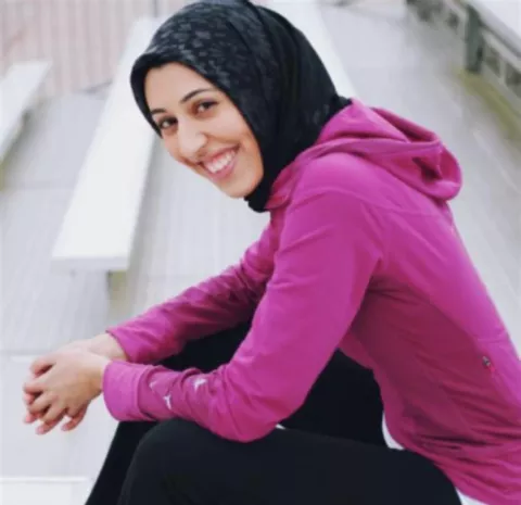 A smiling woman, seated on a bench, wearing a purple hoodie and a black hijab.