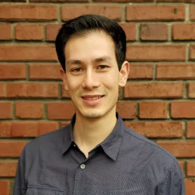 A slightly smiling man with short hair, wearing a dark blue shirt and standing in front of a brick wall.