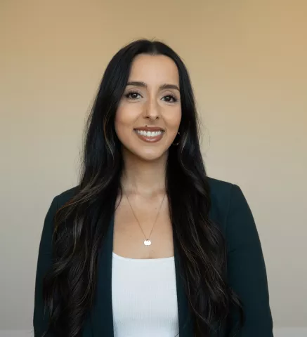 A smiling woman with long dark hair, wearing a white top and a dark green blazer in front of a plain backdrop.