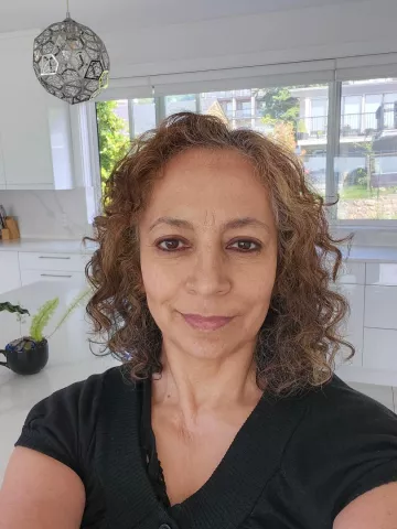 A woman with shoulder-length curly hair, wearing a black shirt, looking into the camera with a faint smile, standing in what may be a kitchen.