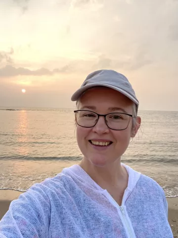 A smiling woman standing in front of a beach sunset, wearing a baseball cap and a white sweater.