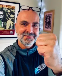 A bald white man with a grey goatee, glasses perched on his head, raising his fist.