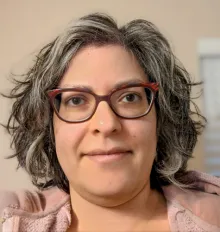 A woman, possibly south Asian, with wavy dark grey hair stares into the camera lens.