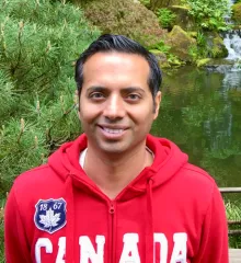 A short-haired man wearing a red hoodie with "Canada" across the front, standing in front of nature.