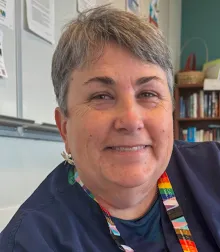 A smiling white woman with short grey hair wearing a navy t-shirt and a rainbow lanyard.