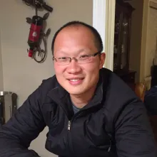 An asian man with close-cropped hair, wearing a black jacket, sitting inside a home.